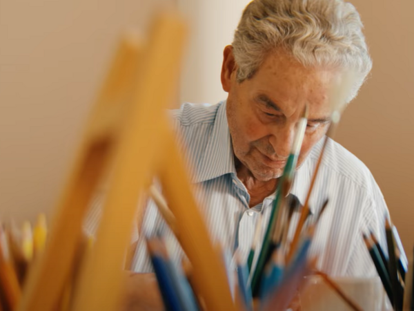 Ein Ehrenamtlicher der Kunsttherapie im Film Gesehen-Fühlen-Leben von Filmemacher Andrew Agbottah über die Menschen im Diakonie Hospiz Wannsee in Berlin sitzt an einem Tisch mit Kunst- und Malutensilien und blickt auf ein Bild Ein Ehrenamtlicher der Kunsttherapie im Film Gesehen-Fühlen-Leben von Filmemacher Andrew Agbottah über die Menschen im Diakonie Hospiz Wannsee in Berlin sitzt an einem Tisch mit Kunst- und Malutensilien und blickt auf ein Bild