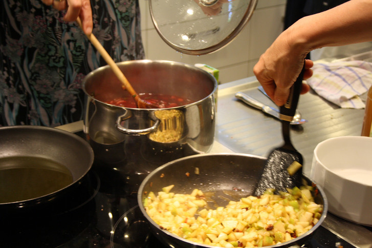 Eine Person rührt Sauce in Topf um und eine andere Person wendet mit Bratenwender Essen in Pfanne Eine Person rührt Sauce in Topf um und eine andere Person wendet mit Bratenwender Essen in Pfanne