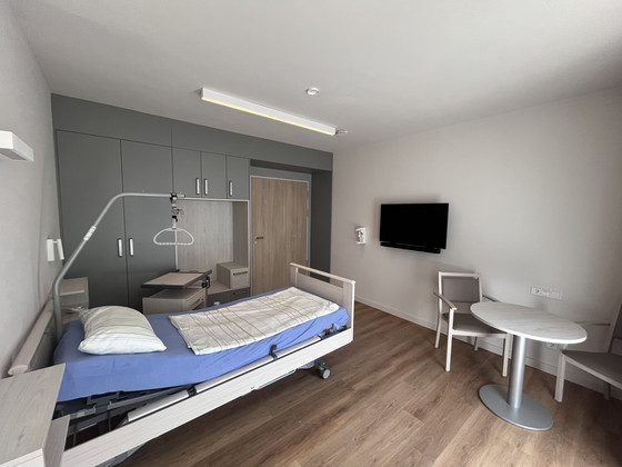 Blick in ein helles Gästezimmer mit einem frisch bezogenen Gästebett, Holzboden, hellen Wandfarben und Monitor an der Wand im Stationären Diakonie Hospiz Wannsee in Berlin-Wannsee. Blick in ein helles Gästezimmer mit einem frisch bezogenen Gästebett, Holzboden, hellen Wandfarben und Monitor an der Wand im Stationären Diakonie Hospiz Wannsee in Berlin-Wannsee.